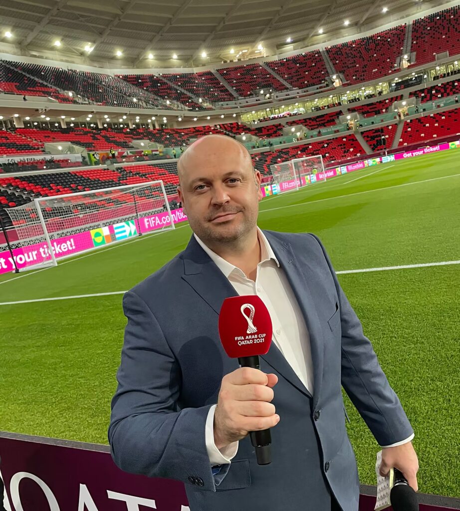 A photo of Alan March in Qatar 2021, holding a microphone with a red windshield in a soccer stadium.
